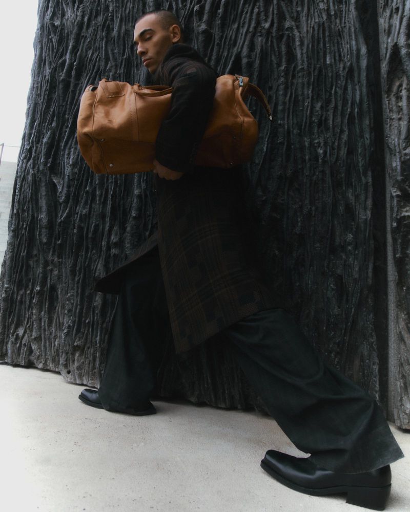 Adult male wearing a dark coat and wide trousers carrying a tan leather duffel bag in front of a textured wall.