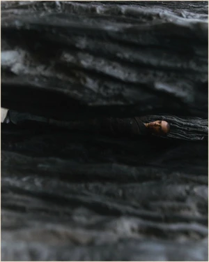 Model lying horizontally inside a narrow opening of dark, textured rock formations in an editorial composition.