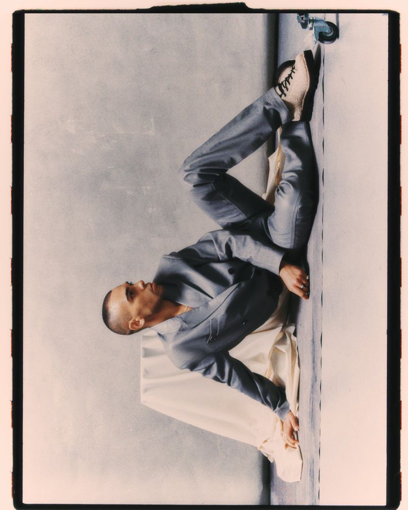 Man with a buzz cut wearing a grey tailored suit reclining on a studio floor against draped neutral fabric.