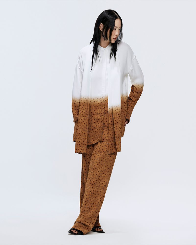 Woman in flowing white-and-brown polka dot shirt and pants stands in photo studio with simple lighting, looking away.