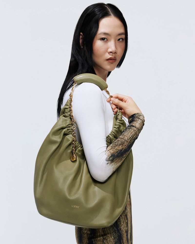 Brunette woman in a white long-sleeve poses with a large, green, gathered-leather Loewe purse with a gold chain and charm accent.