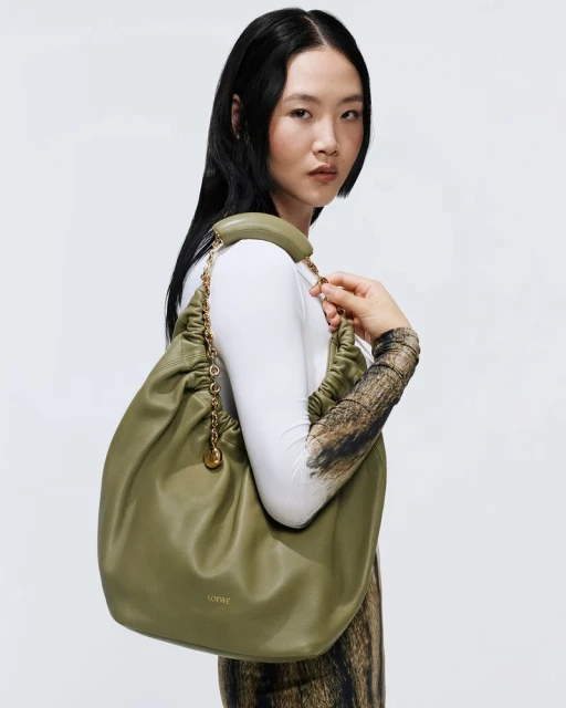 Brunette woman in a white long-sleeve poses with a large, green, gathered-leather Loewe purse with a gold chain and charm accent.