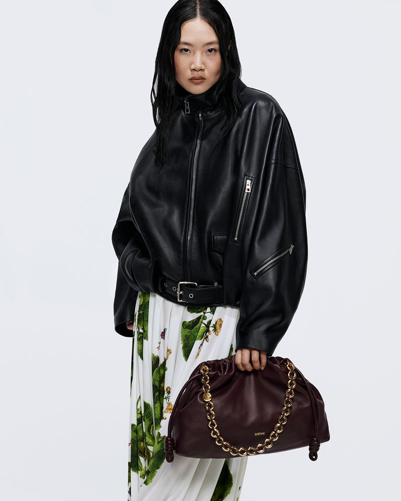 Asian woman in a black leather jacket holds a burgundy handbag with a gold chain. She is wearing a floral skirt in a studio setting.