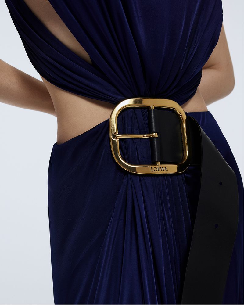 A person wearing a navy dress with a large black and gold belt with the Loewe logo against a solid neutral background. 
