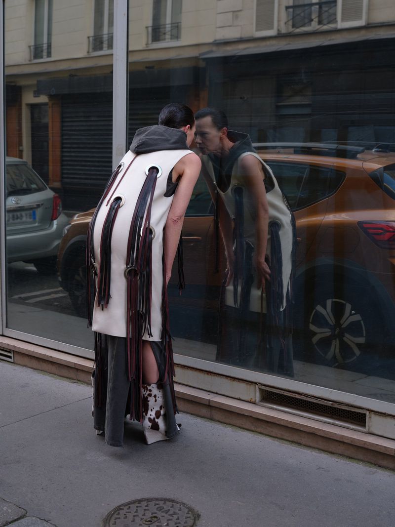 Adult female in a sleeveless white dress with dark brown straps faces her reflection in an urban storefront window in daylight.