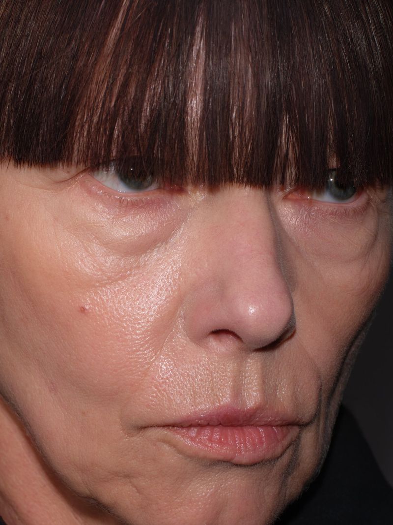Close-up portrait of an adult woman with short brown hair and bangs, fair skin, natural makeup, and a neutral expression against gray.
