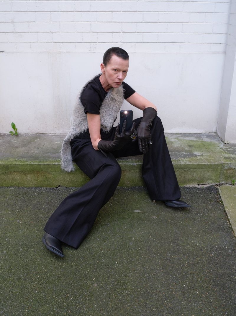 A person with short hair sits on steps outside, wearing a gray vest, black shirt, black pants, and black gloves, holding a cell phone.