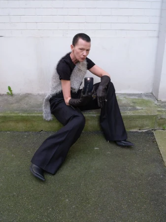 A person with short hair sits on steps outside, wearing a gray vest, black shirt, black pants, and black gloves, holding a cell phone.