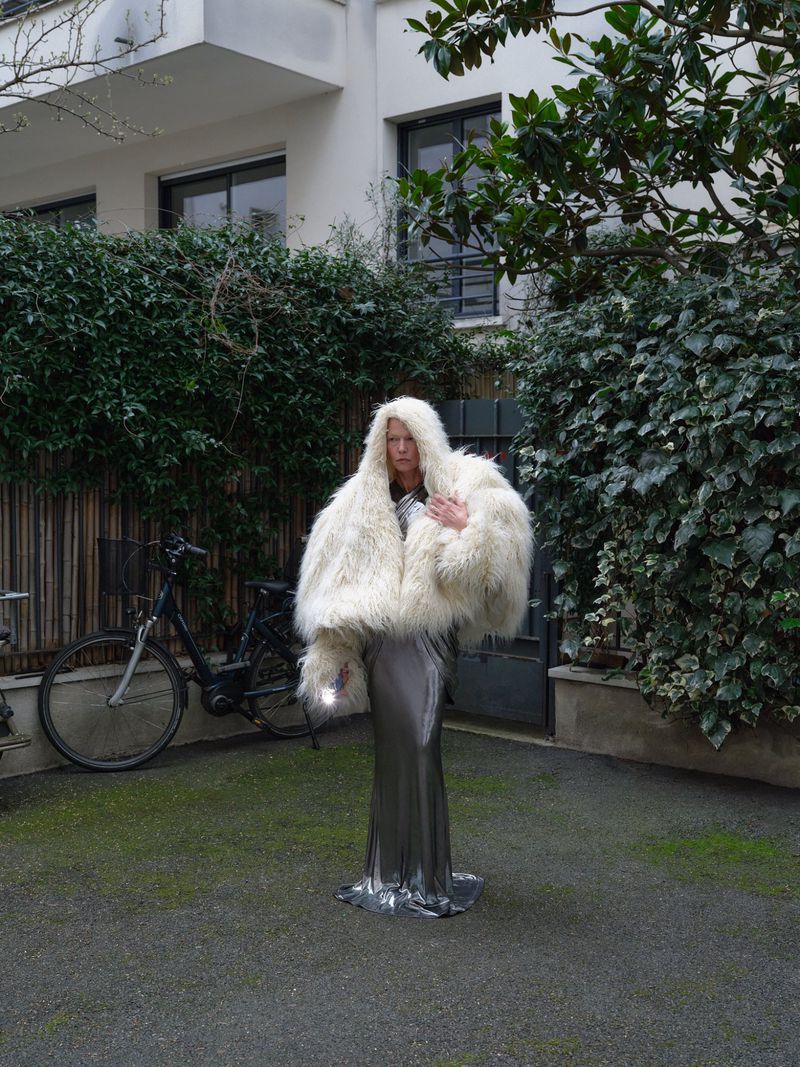 Woman in a large white fur coat and silver dress stands in an outdoor courtyard with green plants and a bicycle in the background.