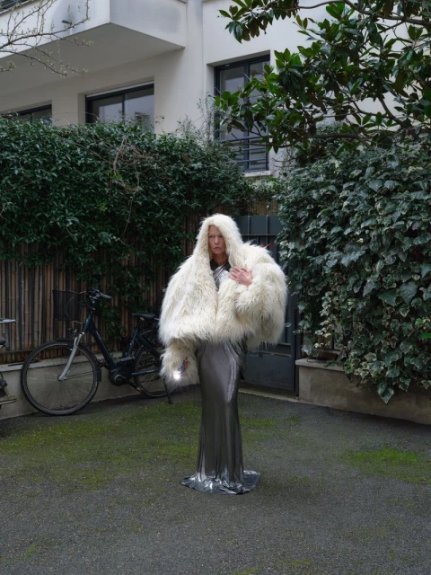Woman in a large white fur coat and silver dress stands in an outdoor courtyard with green plants and a bicycle in the background.