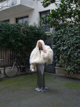 Woman in a large white fur coat and silver dress stands in an outdoor courtyard with green plants and a bicycle in the background.