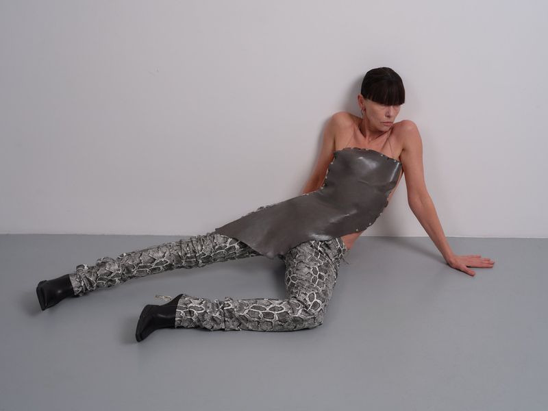 Fashion model poses in a gray strapless top and reptile-print pants against a white wall, shot indoors with gray floor.