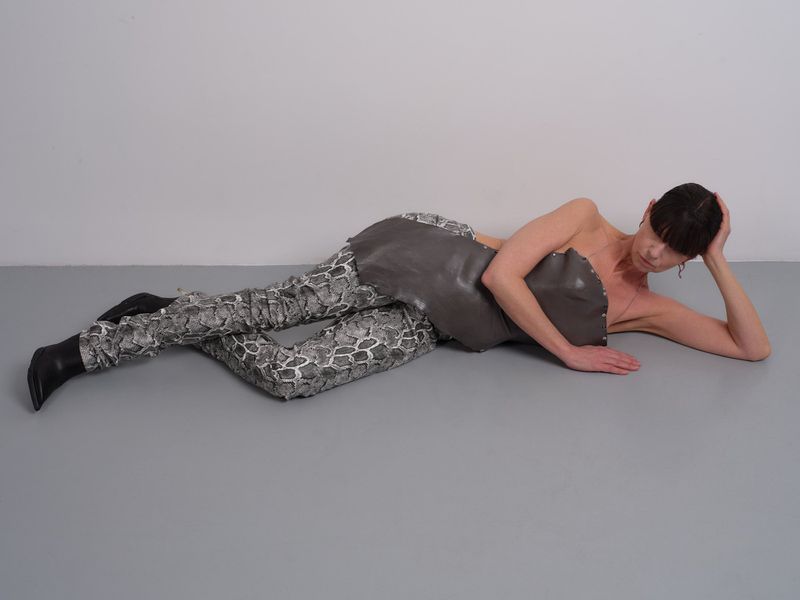 Adult female in grey bodysuit and snake-print pants lies on grey floor in a studio with white wall in background, fashion shot.
