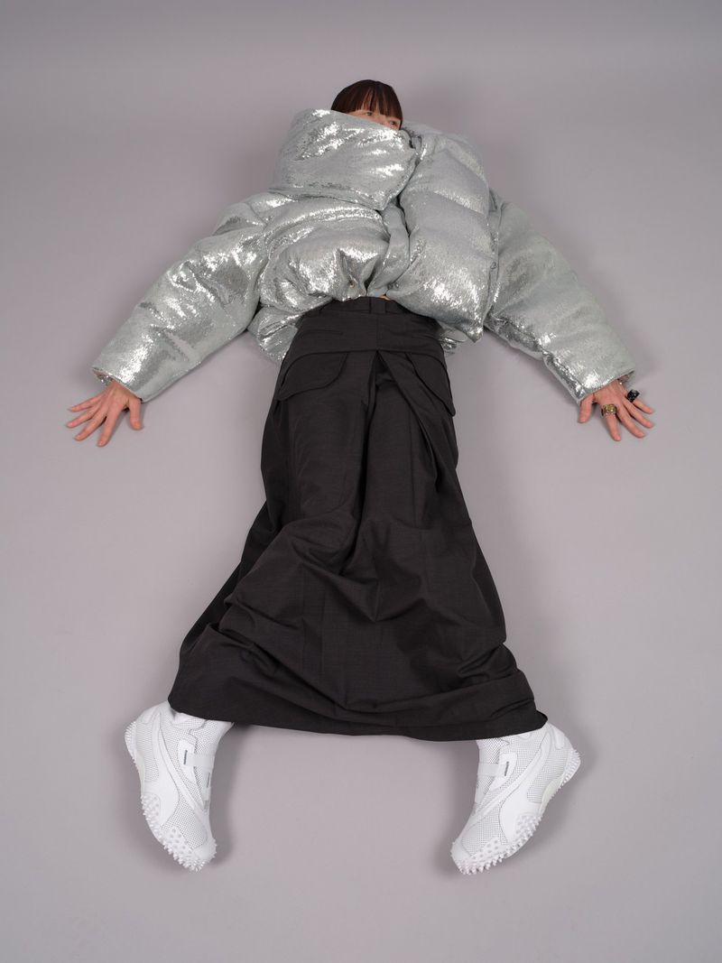 Fashion model wearing a silver puffy coat, black skirt, and white sneakers in a studio shot against a gray background.
