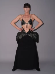Model in dark studded halter top and black sculptural skirt posing against a neutral grey studio backdrop