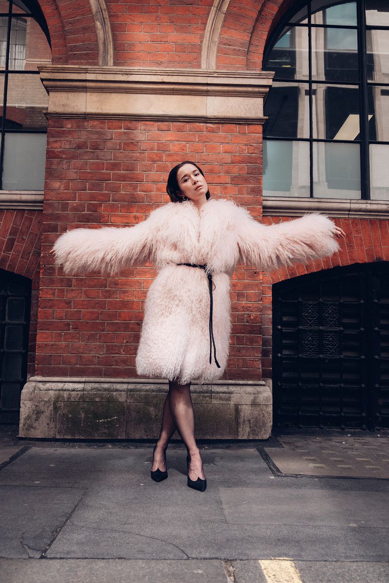 Elisabeth Dulau wearing an oversized pale pink fluffy coat and black heels posing against a red brick building exterior.