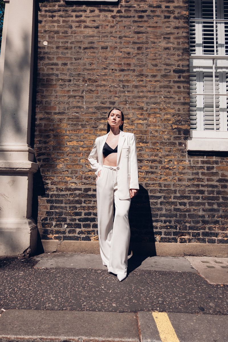 A woman in a white suit poses against a brick wall in an urban setting, showcasing fashion-forward style and natural sunlight.