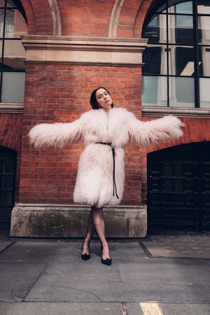 Elisabeth Dulau wearing an oversized pale pink fluffy coat and black heels posing against a red brick building exterior.