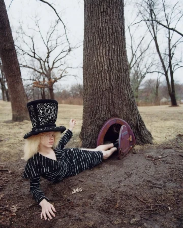 An adult female lies near a tree with a miniature door wearing a black and white striped dress and a large decorative top hat outdoors.