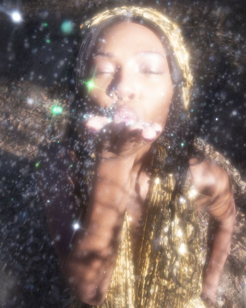 Woman in golden headscarf blows glitter towards camera in forest with bright sun and bokeh effects, creating a dreamy atmosphere.
