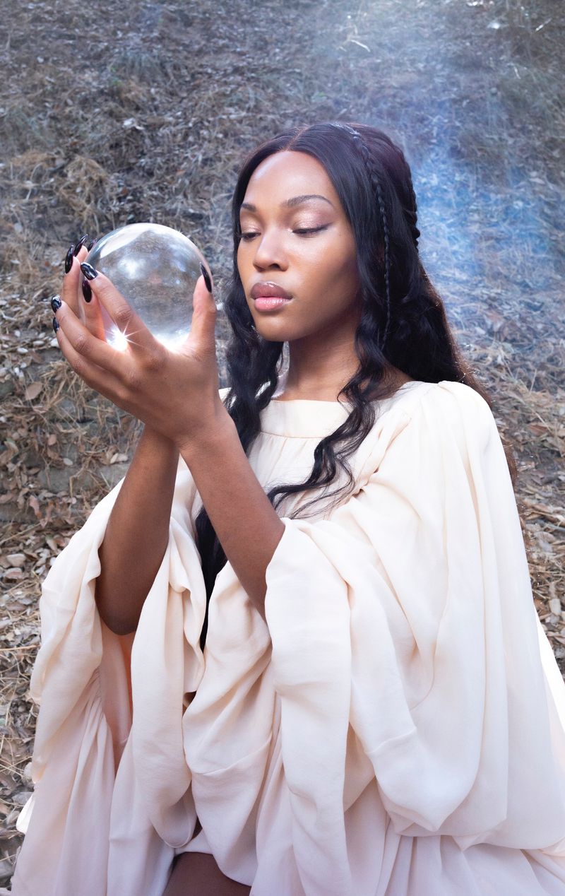 Elegant woman in flowing gown holding a glass orb in a dreamy outdoor setting, mystical aesthetic, soft focus, creating a serene and ethereal image.
