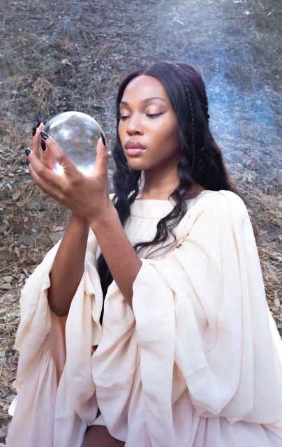 Elegant woman in flowing gown holding a glass orb in a dreamy outdoor setting, mystical aesthetic, soft focus, creating a serene and ethereal image.