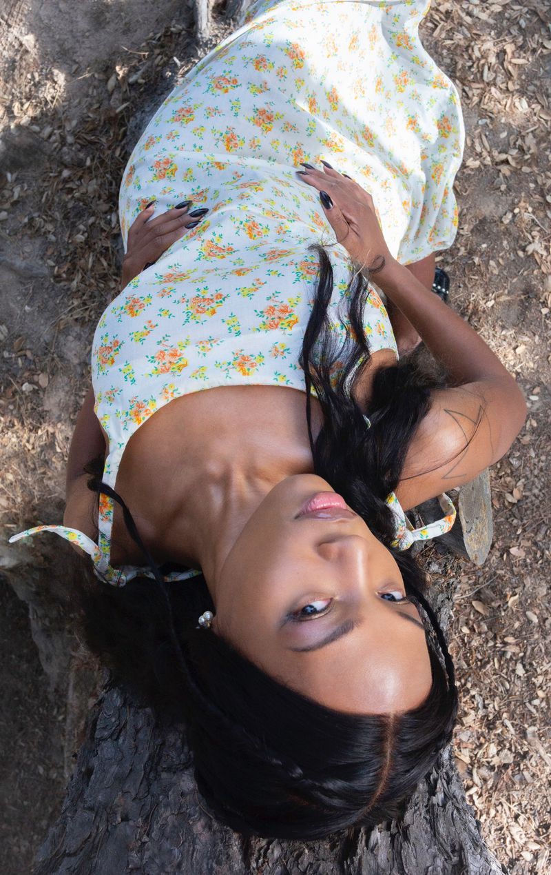 A woman in a floral dress lies on a log outdoors, looking up at the camera with dark hair and black nails. Natural light.