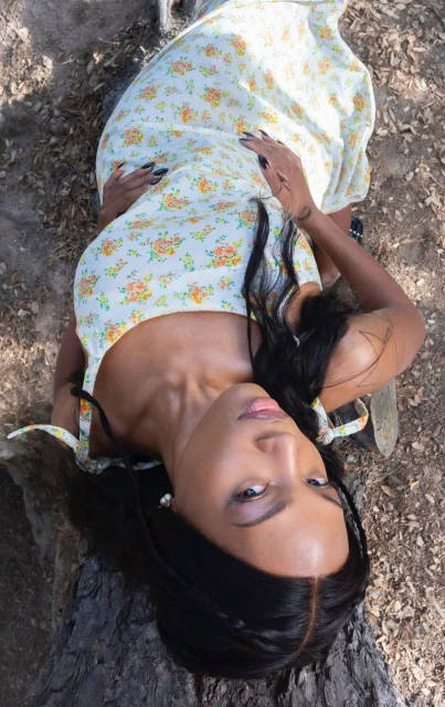 A woman in a floral dress lies on a log outdoors, looking up at the camera with dark hair and black nails. Natural light.