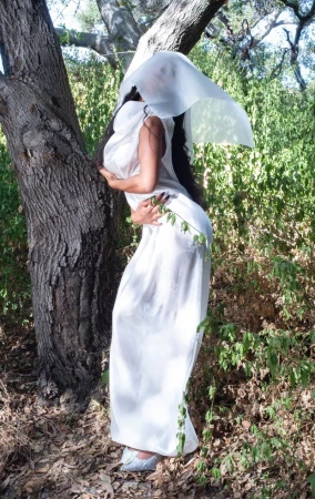 A woman draped in white fabric poses outdoors, near a tree, with greenery visible in the background, creating an ethereal, fashion-inspired image.