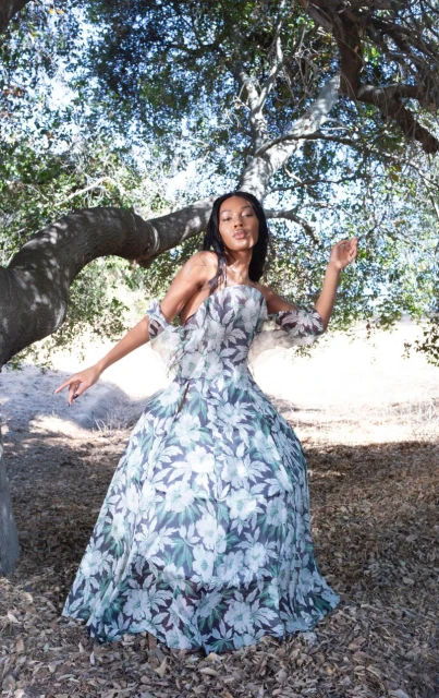 Woman in an elegant floral print gown stands under a tree with dappled lighting in an outdoor setting with dried leaves on the ground.