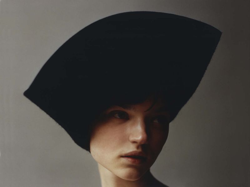 A headshot of an adult female with fair skin wearing a large black hat, looking away in a brightly lit indoor photo studio.
