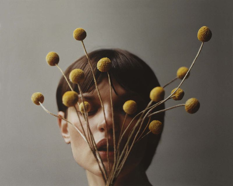Woman with short brown hair partially obscured by a bouquet of round, yellow Billy Buttons flowers against a solid grey backdrop.