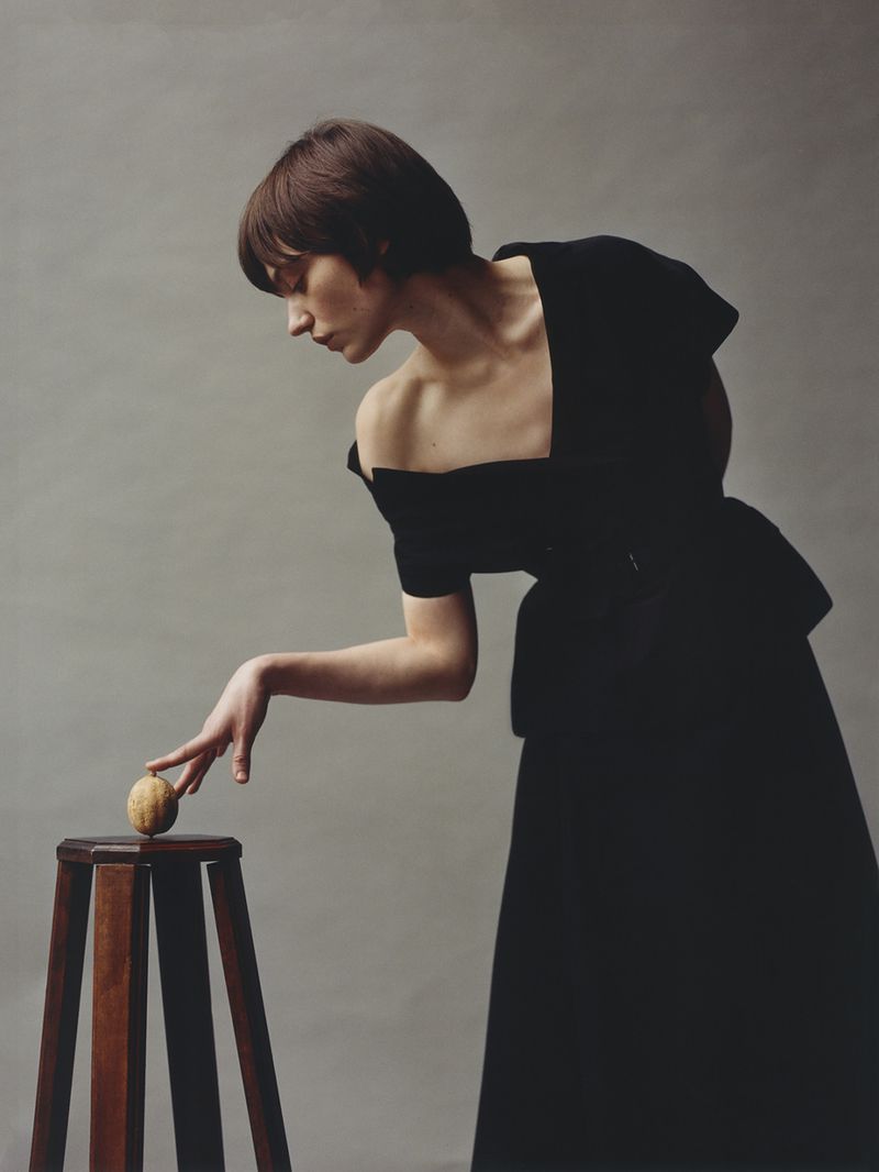A slender woman in a black dress poses with a walnut on a wooden stool, set against a grey backdrop in a studio setting.