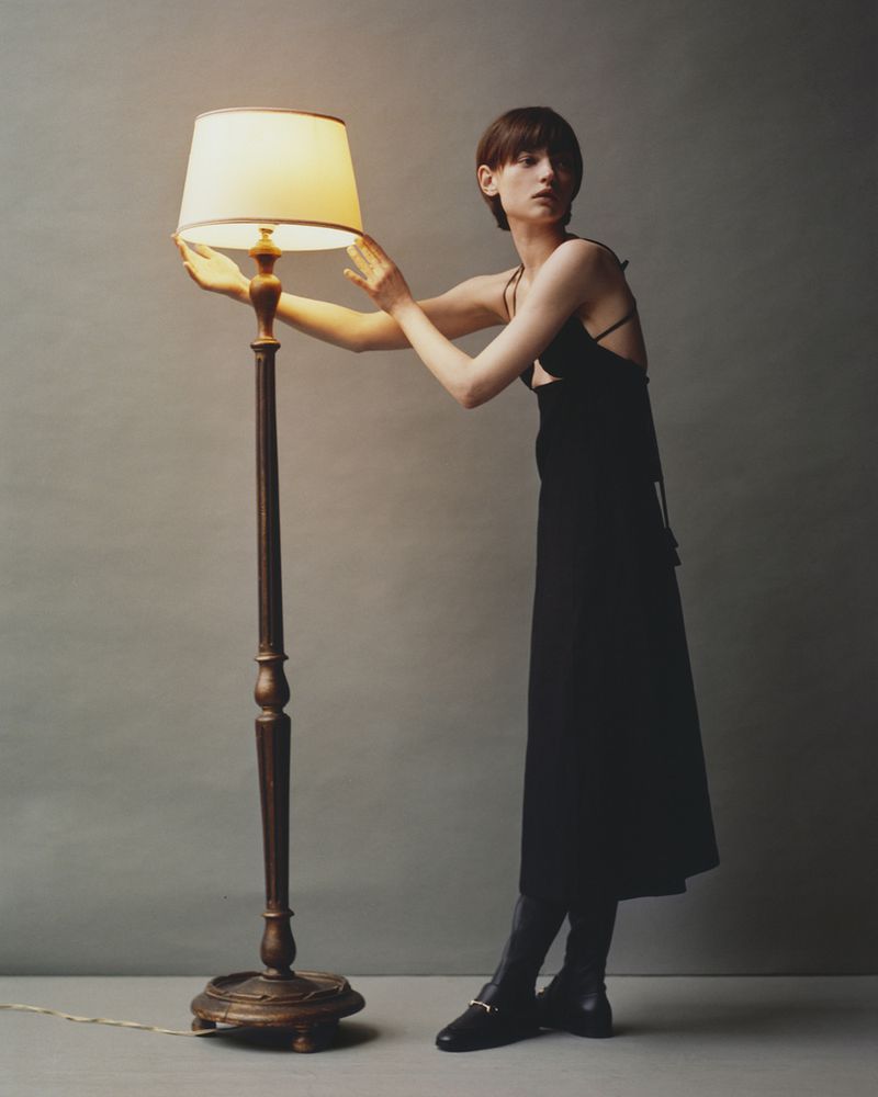 Woman in black dress poses near a floor lamp. The lamp is lit, casting a warm glow. Dark boots are visible against the neutral background.