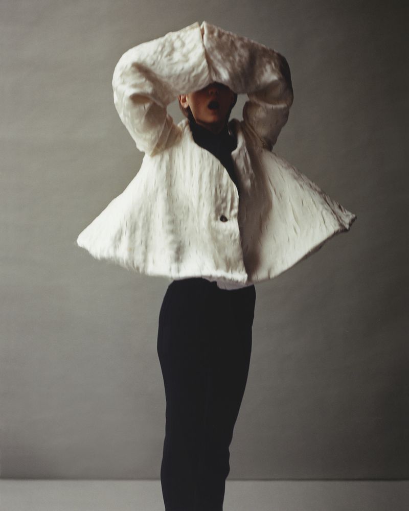 Person in black pants with sculptural textured cream-colored jacket posing against a neutral backdrop in studio lighting.