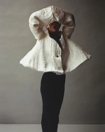 Person in black pants with sculptural textured cream-colored jacket posing against a neutral backdrop in studio lighting.