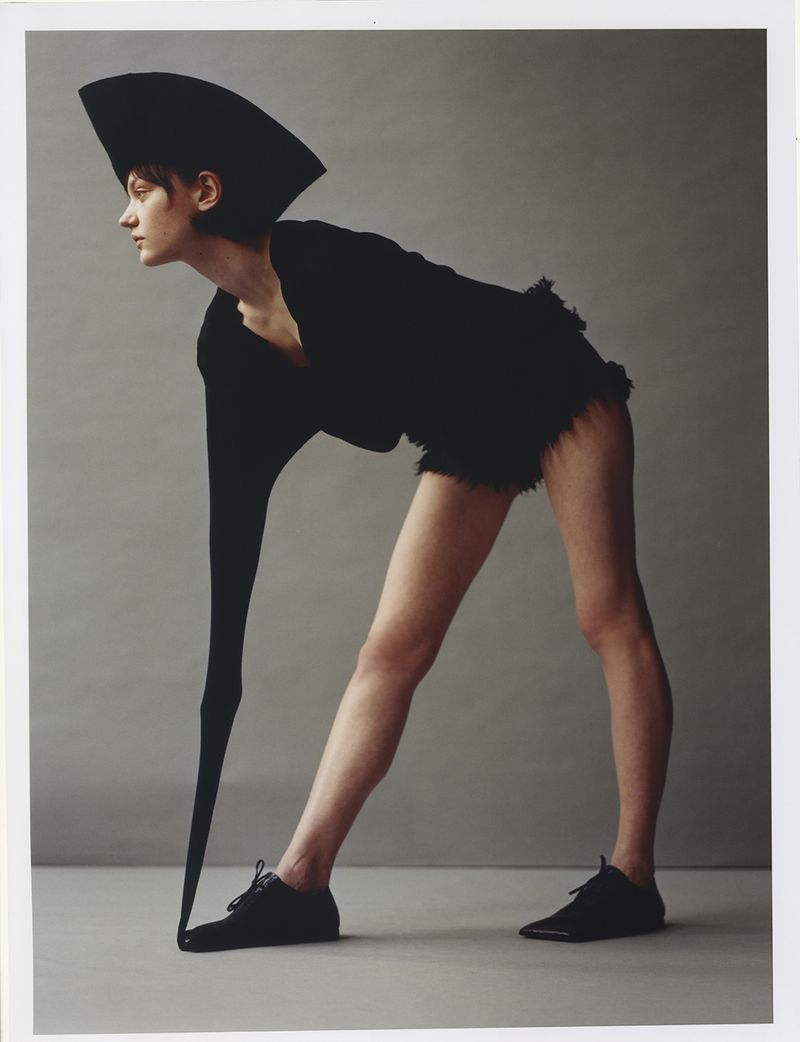 Fashionable adult female model in a sculptural black hat and matching top with shorts, posing in front of a grey backdrop.