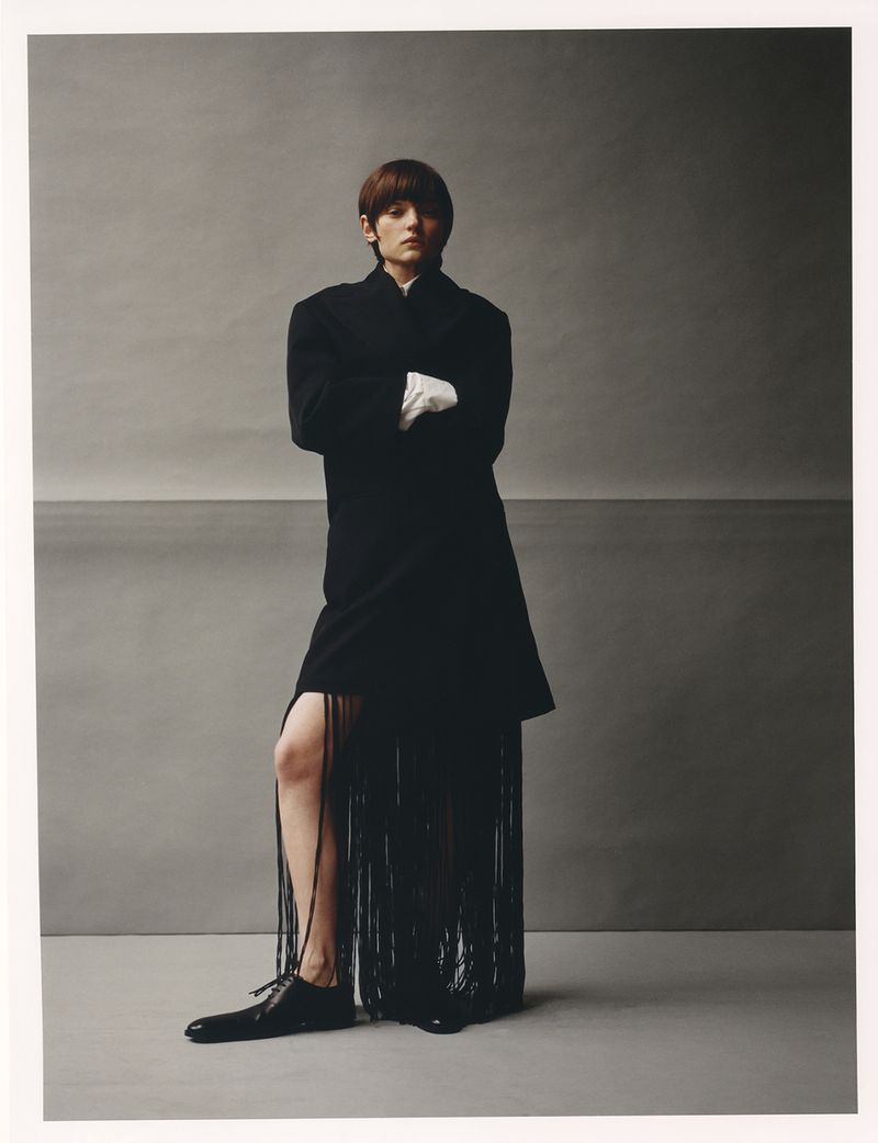Woman poses in a black coat with fringe and white gloves against a grey backdrop in a studio setting, fashion-forward and stylish.