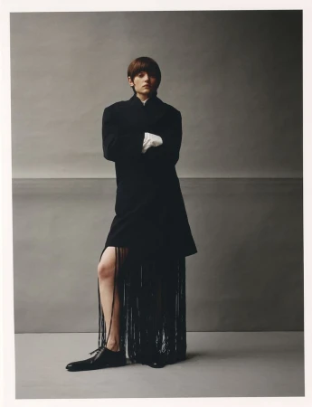 Woman poses in a black coat with fringe and white gloves against a grey backdrop in a studio setting, fashion-forward and stylish.