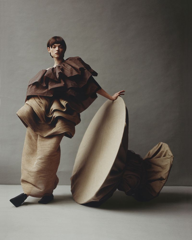 Woman in brown tiered ruffle garment poses next to a large tan round object against a grey background in a fashion studio shot.