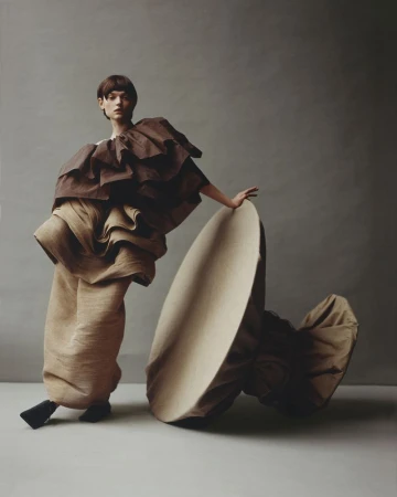 Woman in brown tiered ruffle garment poses next to a large tan round object against a grey background in a fashion studio shot.