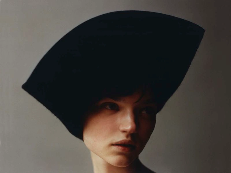 Model Pasha Harulia wearing a large sculptural black curved hat against a muted grey background.