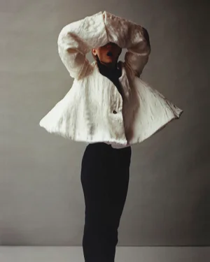 Model posing in a sculptural off-white textured cape jacket and black trousers against a neutral grey studio backdrop.