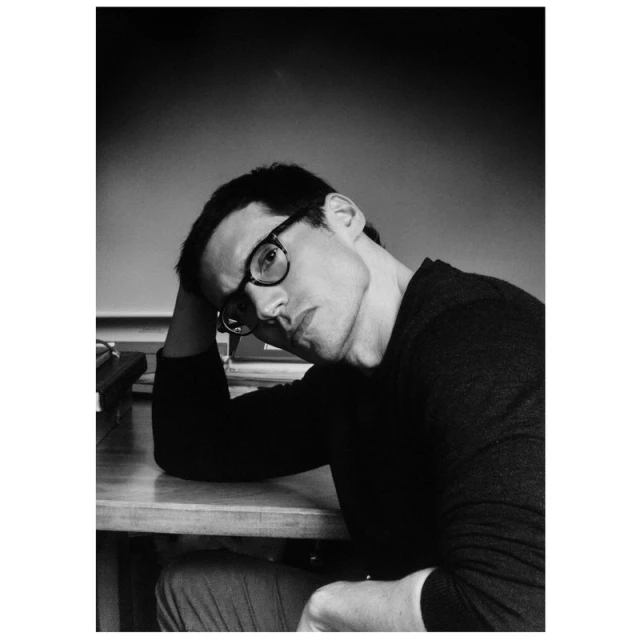 Black and white portrait of a man wearing glasses sitting at a table with his head resting on his hand in an indoor setting.
