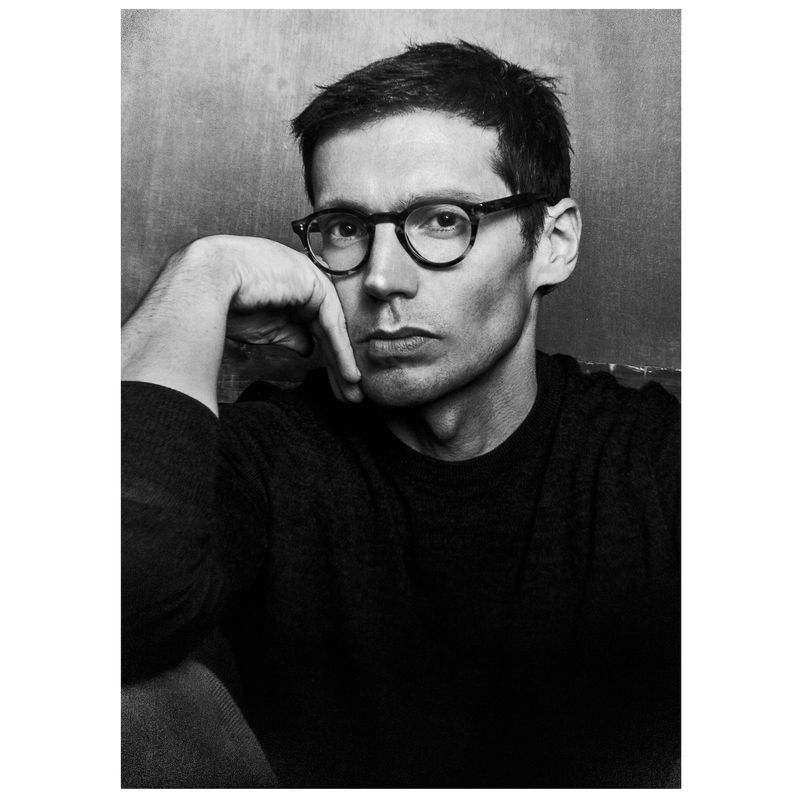 Black and white portrait of designer Erdem Moralioglu wearing glasses and resting his face on his hand against a textured wall.