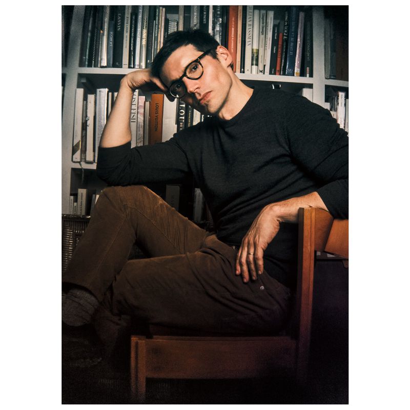 An adult male sits in a wooden chair with books in the background, wearing glasses, a dark shirt, and brown pants in an indoor setting.