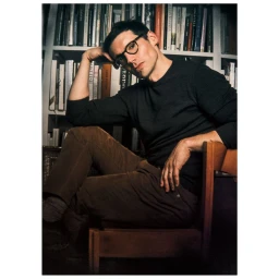 Man in round glasses and dark sweater seated on a wooden chair against a bookshelf filled with books