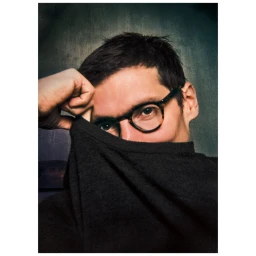 Man wearing round eyeglasses holding a dark knit sweater up over the lower half of his face against a textured background