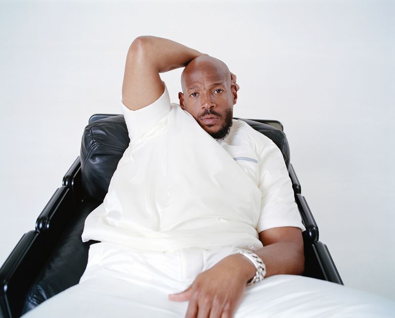 Adult Black male in a light outfit sitting in a black leather chair against a white background in a studio setting with casual pose.