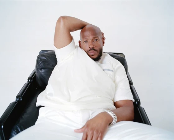 Adult Black male in a light outfit sitting in a black leather chair against a white background in a studio setting with casual pose.